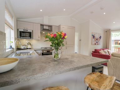 A kitchen with flowers in a vase on a countertop and a living area with a red sofa and artwork on the wall at Kirkstone Lodge in Limefitt Park near Windermere