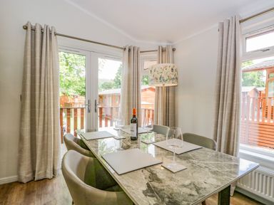 A dining area with a marble table set with placemats wine glasses and a bottle of wine near windows with beige curtains at Kirkstone Lodge in Limefitt Park near Windermere