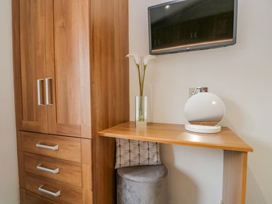 A corner with a wooden cabinet, a wall-mounted television, a wooden desk with a round mirror and a vase with flowers at Kirkstone Lodge in Limefitt Park near Windermere