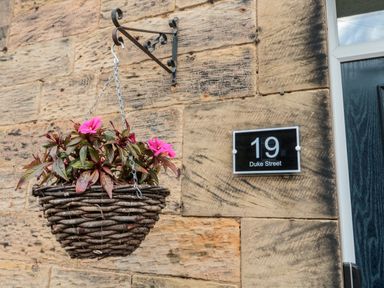 A hanging basket with pink flowers on a stone wall next to a house number sign reading 19 Duke Street at 19 Duke Street in Alnwick