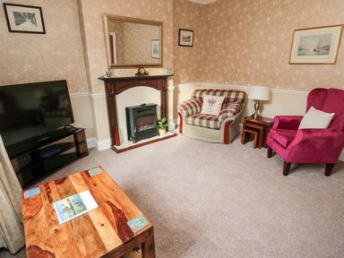 A living room with a television on a stand a wooden coffee table a fireplace with a mirror above a plaid armchair a red armchair and a lamp on nested tables at 19 Duke Street Alnwick