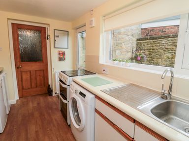 A kitchen with a washing machine stove and sink under a window showing an outside stone wall at 19 Duke Street in Alnwick