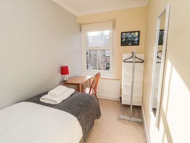 A small bedroom with a single bed, a wooden desk and chair, a red lamp, a window, a standing mirror, and a white storage unit at 19 Duke Street in Alnwick