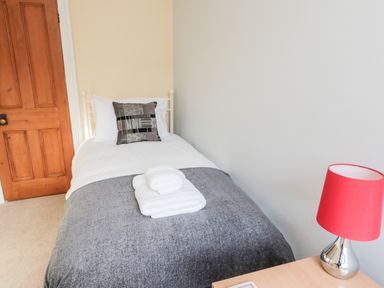A single bed with folded towels a patterned cushion a wooden door and a bedside table with a red lamp at 19 Duke Street in Alnwick