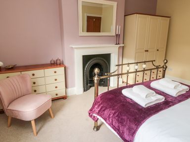 A bedroom with a metal bed, white and purple bedding, a pink chair, cream wardrobe, chest of drawers, and a fireplace with a mirror at 19 Duke Street in Alnwick