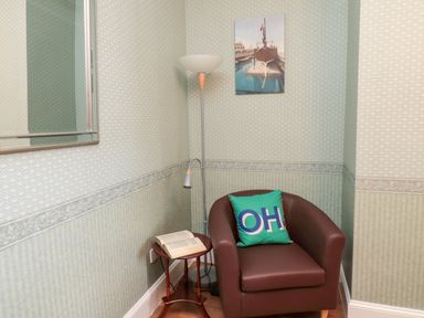 A corner with a brown armchair with a green pillow reading OH a small wooden side table with an open book and a floor lamp at 19 Duke Street in Alnwick