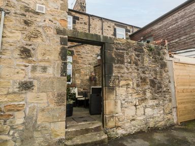 An outdoor stone walled patio area with steps leading to a seating area with chairs and a hanging flower pot at 19 Duke Street in Alnwick