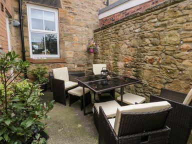 An outdoor seating area with a glass table and six cushioned chairs surrounded by stone walls at 19 Duke Street in Alnwick