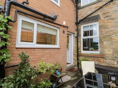 A small outdoor patio area with patio furniture a table and chair plants and the back doors and windows of two adjoining brick and stone buildings at 19 Duke Street Alnwick