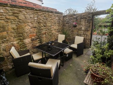 An outdoor patio with black glass table and wicker chairs with cushions next to a stone wall at 19 Duke Street in Alnwick