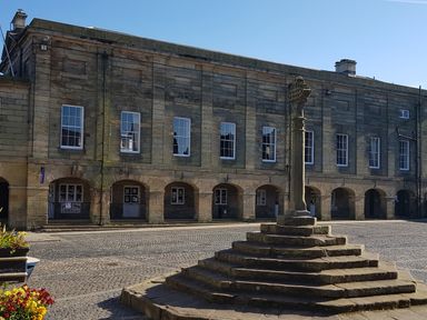 A stone market cross on stepped base in a paved open square in front of a building with arched entrances and rectangular windows at 19 Duke Street in Alnwick