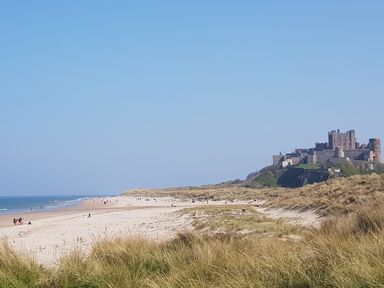A sandy beach with people walking near the water and a large stone castle on a hill overlooking the beach at 19 Duke Street in Alnwick