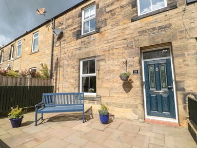 A blue bench and two flower pots outside a stone house with a blue door at 19 Duke Street in Alnwick