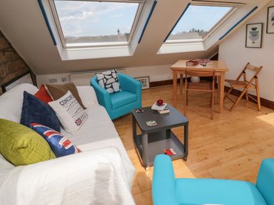 A living room with a white sofa with pillows two blue armchairs a small black coffee table and a wooden table with chairs under skylight windows at 19 Duke Street in Alnwick