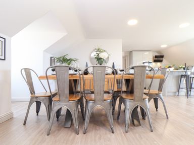 A dining area with a wooden table and metal chairs in a bright room with kitchen in the background at Penthouse 23 in Newquay
