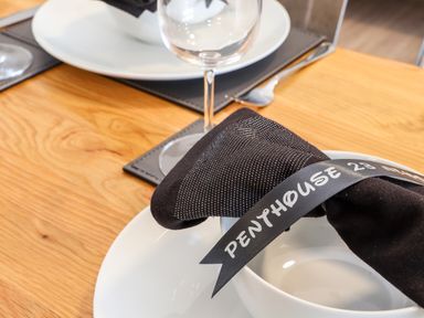 A close up of a dining table setup with a white plate a black napkin with a ribbon that says Penthouse 23 and a wine glass on a wooden table at Penthouse 23 in Newquay