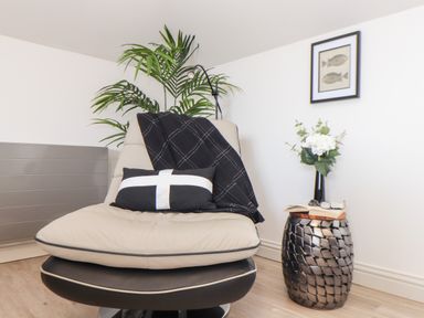 A beige chair with a black and white pillow and a black blanket in a room with a plant a side table with a book glasses and flowers and a picture of fish on the wall at Penthouse 23 in Newquay