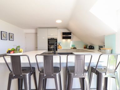 A kitchen with four metal bar stools and a bowl of fruit on a white countertop at Penthouse 23 in Newquay