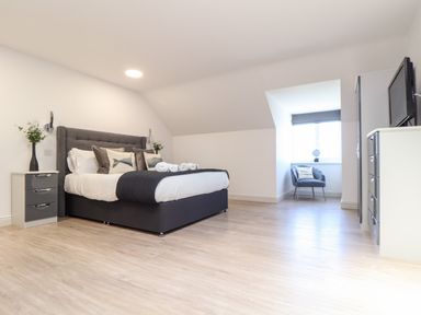 A bedroom with a double bed and bedside table with a vase and flowers a chair by a window and a chest of drawers with a tv on top at Penthouse 23 in Newquay