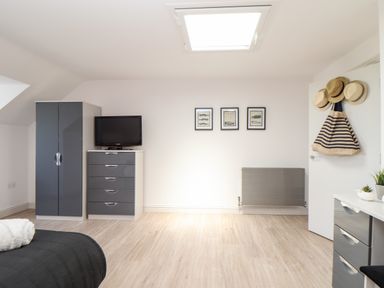 A bedroom with a gray wardrobe and chest of drawers with a television a bed corner is visible and hats hanging on a door at Penthouse 23 in Newquay