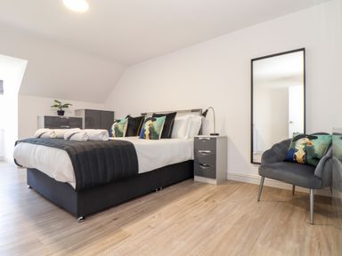 A bedroom with a double bed with white and black bedding and seagull themed cushions with a grey chair holding seagull themed cushions a tall mirror and grey drawers at Penthouse 23 in Newquay