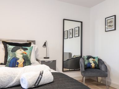 A bedroom with a bed and a gray chair both with seagull printed cushions a bedside table with a lamp and a full length mirror on the wall at Penthouse 23 in Newquay