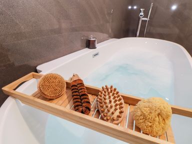A bathtub filled with water and foam with a wooden bath tray holding three brushes and a natural sponge at Penthouse 23 in Newquay