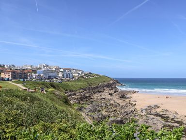 A coastal cliff with rocks and green vegetation overlooking a sandy beach and ocean with buildings in the background at Penthouse 23 in Newquay