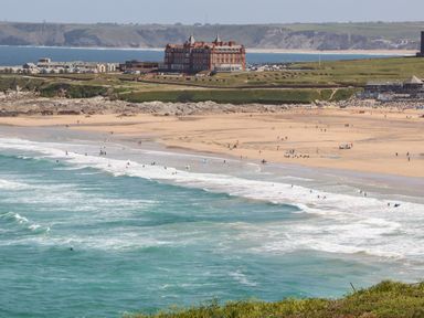 A beach with people surfing and walking near the ocean and a large red building on the cliff at Penthouse 23 in Newquay