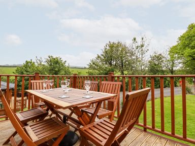 An outdoor wooden deck with a wooden table and six chairs with wine glasses on the table at 23 Bossiney Bay in Tintagel