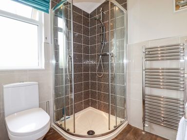 A bathroom with a corner shower, a toilet, a window with a blue blind, and a chrome towel radiator at 23 Bossiney Bay in Tintagel