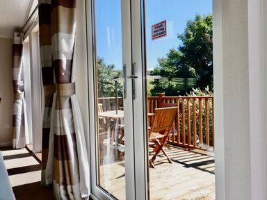 Glass double doors with curtains leading to a wooden deck with table and chairs surrounded by trees at 23 Bossiney Bay in Tintagel