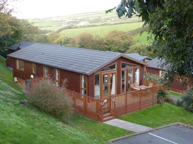 A wooden cabin with a deck and outdoor furniture on a grassy slope with fields in the background at 23 Bossiney Bay in Tintagel