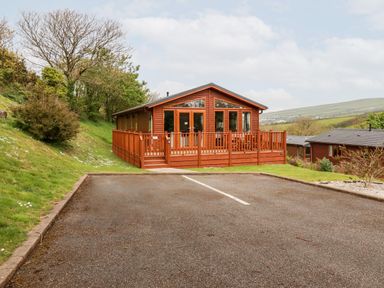 A wooden cabin with a deck and parking spaces surrounded by grassy hills in Tintagel