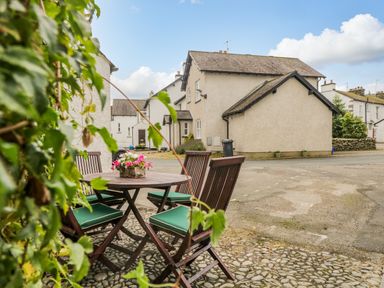 An outdoor seating area with a round wooden table and four chairs with green cushions on a cobblestone surface with white houses in the background at Columbine Cottage in Hawkshead
