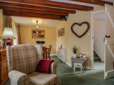 A living room with a plaid armchair a red blanket wooden beams a dining table with chairs and a heart-shaped wall decoration at Columbine Cottage in Hawkshead
