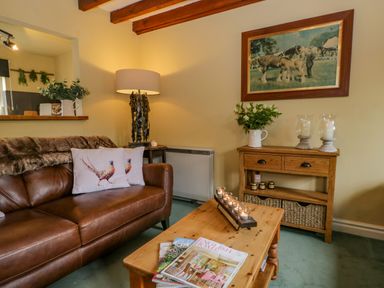A living room with a brown leather sofa and pillows near a wooden coffee table and side table with a lamp and flowers at Columbine Cottage in Hawkshead