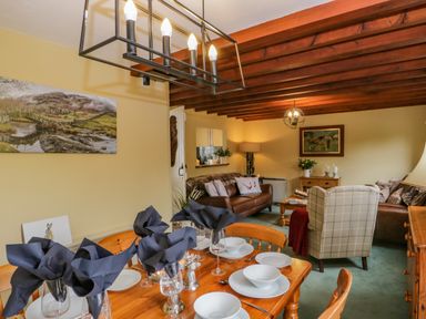 A dining table set with white dishes and black napkins next to a living room with sofas and wooden ceiling beams at Columbine Cottage in Hawkshead
