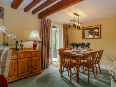 A dining room with a wooden table set for six with black napkins and wooden chairs beside a wooden cabinet with a lamp and bottles at Columbine Cottage in Hawkshead