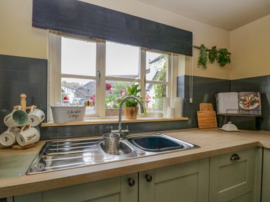 A kitchen sink under a window with cups on a rack and a cookbook open on the counter at Columbine Cottage in Hawkshead