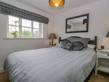 A bedroom with a bed with grey bedding two bedside tables with lamps a window with blinds and a mirror on the wall at Columbine Cottage in Hawkshead