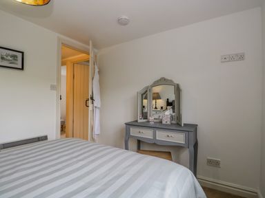 A bedroom with a striped bedspread a dressing table with a tri-fold mirror and an open door showing a hallway at Columbine Cottage in Hawkshead