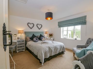 A bedroom with a bed with cushions and a tray bedside tables with lamps heart shaped wall decor armchair and window at Columbine Cottage in Hawkshead