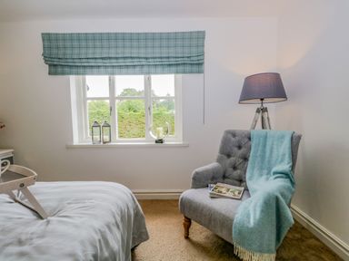 A bedroom corner with a window, two lanterns on the windowsill, a grey chair with a blue throw and magazines, and a floor lamp at Columbine Cottage in Hawkshead