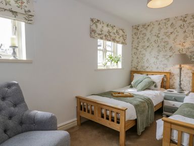 A bedroom with two single wooden beds a grey armchair floral wallpaper on one wall and a bedside table with a lamp at Columbine Cottage in Hawkshead