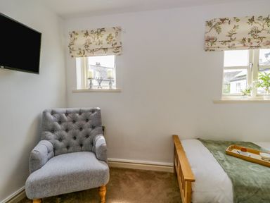 A bedroom with a gray upholstered armchair a wall-mounted television two windows with floral curtains and a bed with a green throw and a wooden tray at Columbine Cottage in Hawkshead