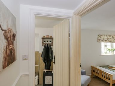 A hallway with a painting of a highland cow leading to a bathroom with towels and a laundry bag and a bedroom with a wooden bed and a window at Columbine Cottage in Hawkshead