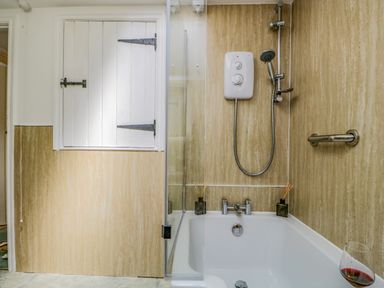 A bathroom with a bathtub with a shower unit and white wooden window shutters at Columbine Cottage in Hawkshead