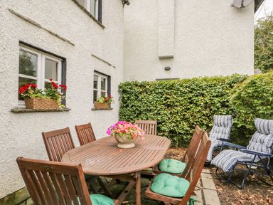 An outdoor patio area with a wooden dining table and chairs with green seat cushions a flower pot on the table two window boxes with red flowers mounted on white roughcast walls two striped reclining chairs and a green hedge