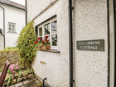 Exterior wall of a cottage with a window box of red flowers and a sign saying Columbine Cottage in Hawkshead